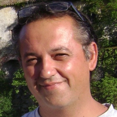 Ritratto di uomo di mezza età sorridente con occhiali sulla testa, all'aperto con vegetazione e muro di pietra.