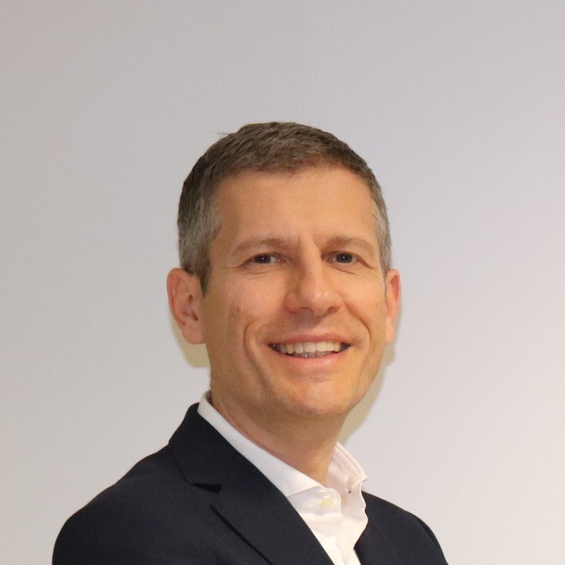 Primo piano di uomo sorridente in giacca su sfondo chiaro.