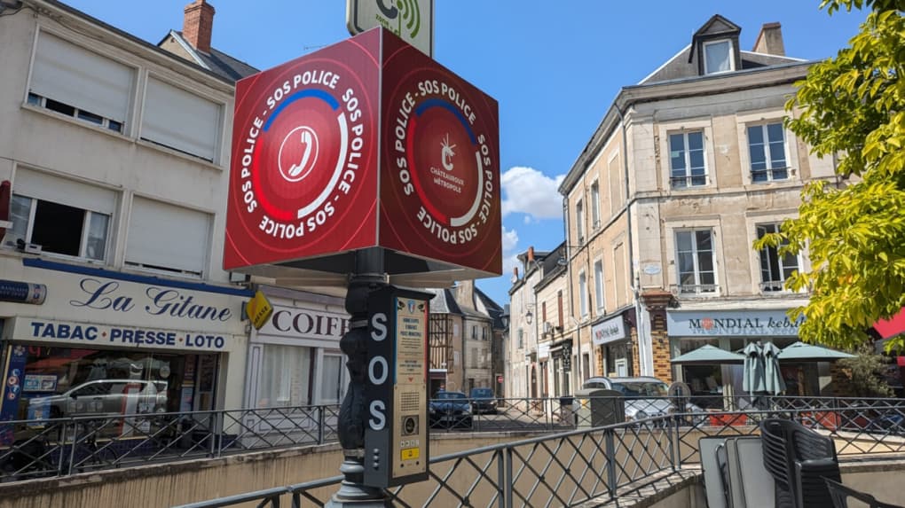 Red SOS Police intercom with phone icon in a French town square, historic buildings, shops.