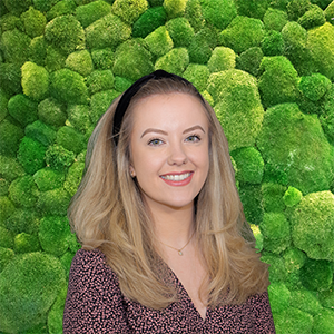 Smiling blonde woman in dark patterned top, black headband, against a vibrant green moss wall.