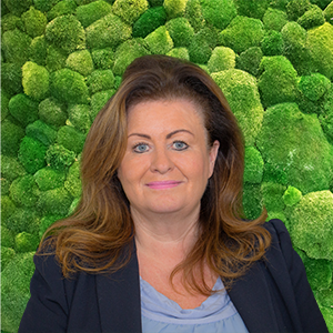 Headshot of a smiling professional woman with brown hair and a dark blazer, set against a green moss wall background.
