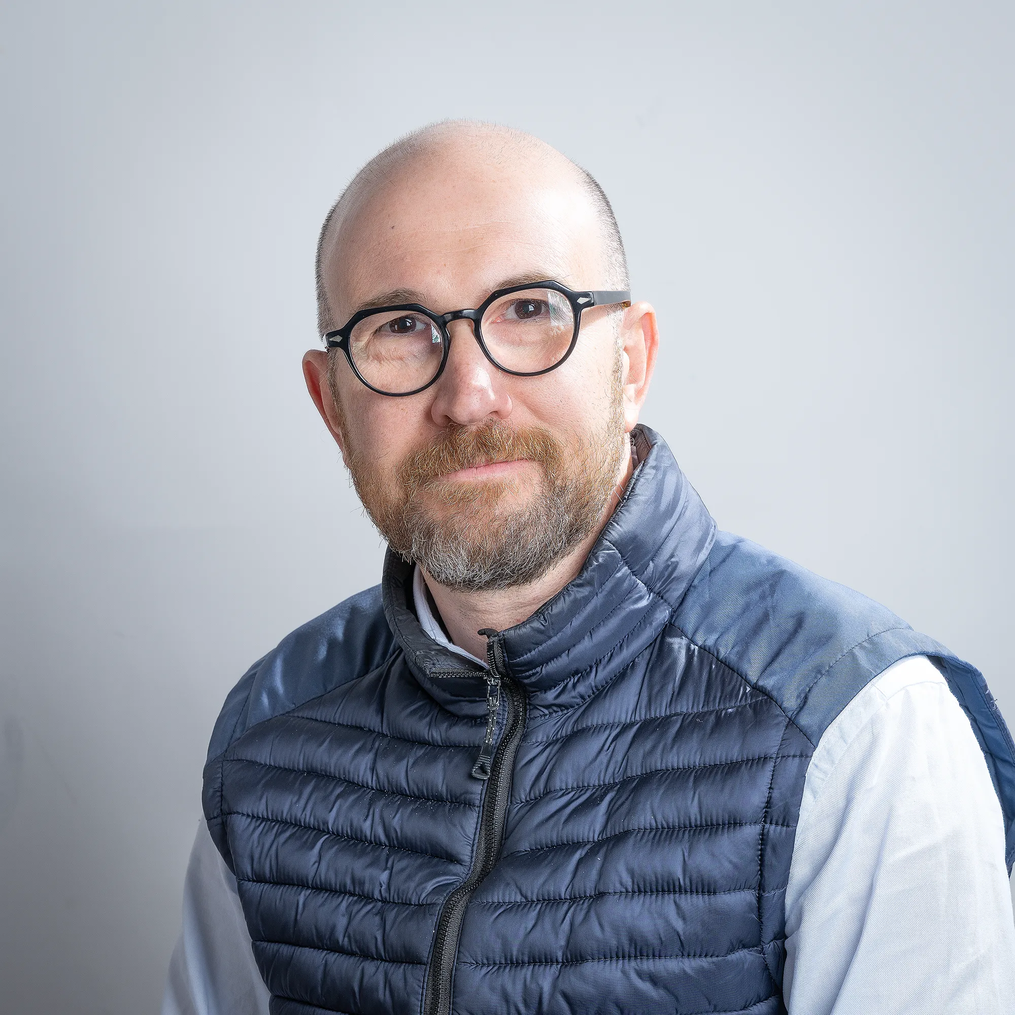 Ritratto di un uomo calvo con barba e occhiali, indossa un gilet blu e guarda verso la telecamera.