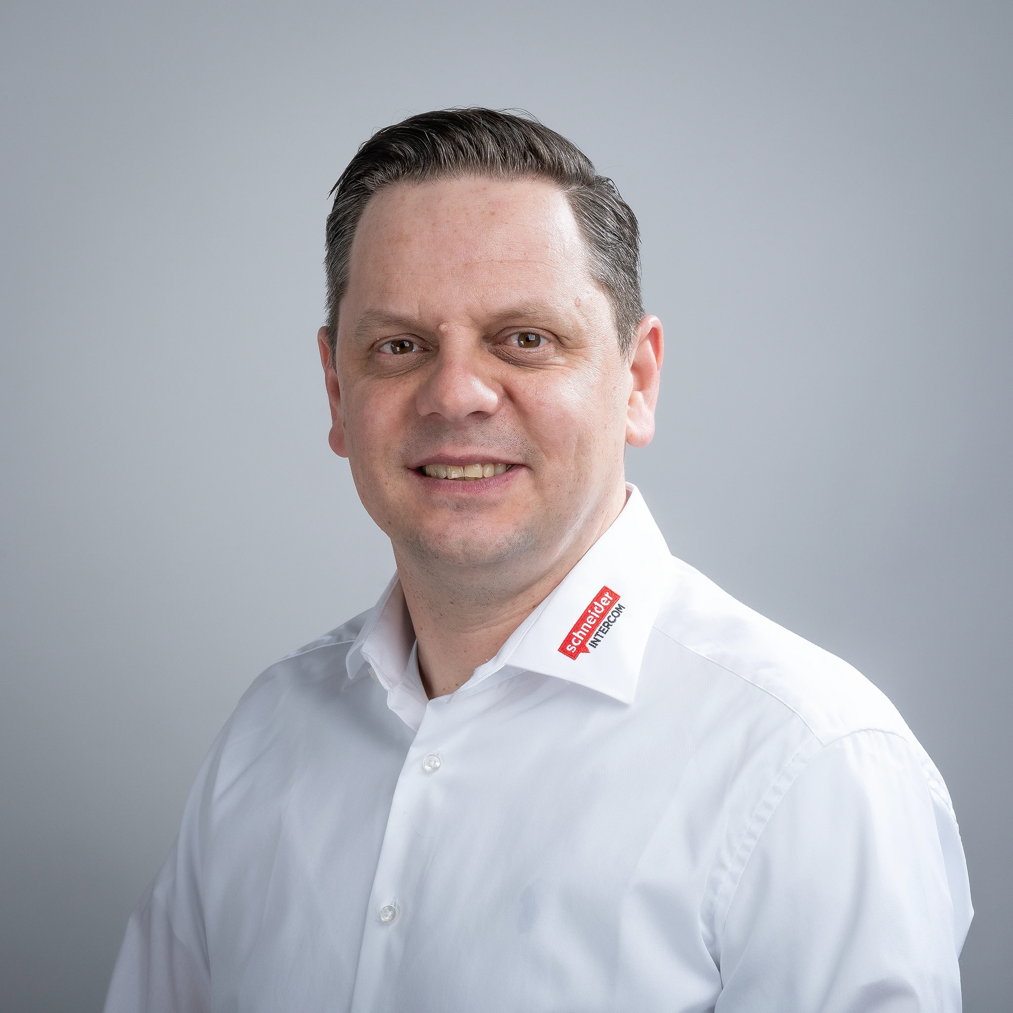 Mann in weißem Hemd mit Schneider Intercom Logo lächelt. Professionelles Portrait zu Kommunikationssystemen.