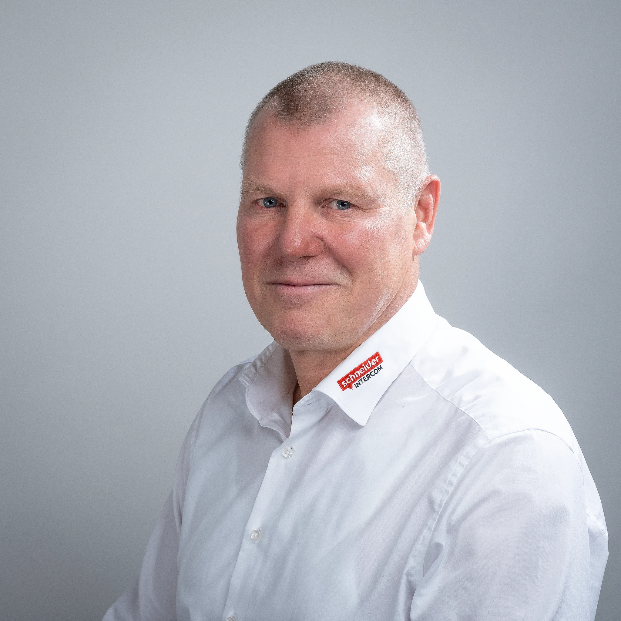 Ein Mann von Schneider Intercom in weißem Hemd lächelt vor grauem Hintergrund.