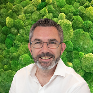 Smiling man with glasses and beard in a white shirt, against a green moss wall background.