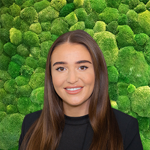 Headshot of a smiling young woman with long brown hair, wearing black, against a green moss wall.