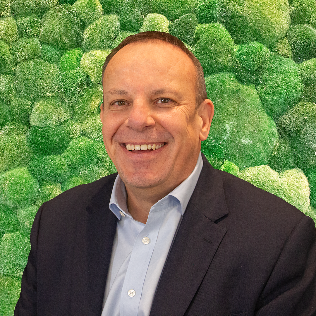 Smiling man in dark suit and light blue shirt against a green moss wall background. Professional headshot.