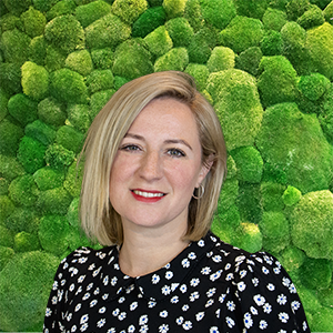 Blonde woman smiling, professional headshot against a green moss wall.