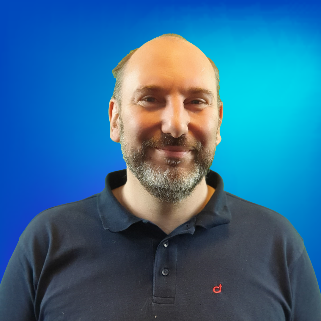 Un homme barbu souriant en polo foncé devant un fond bleu.