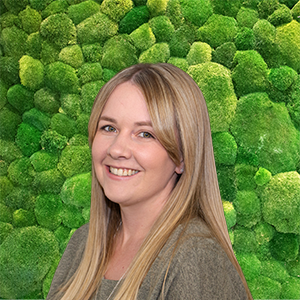 Smiling woman with long blonde hair in front of a green moss wall, representing our friendly team.