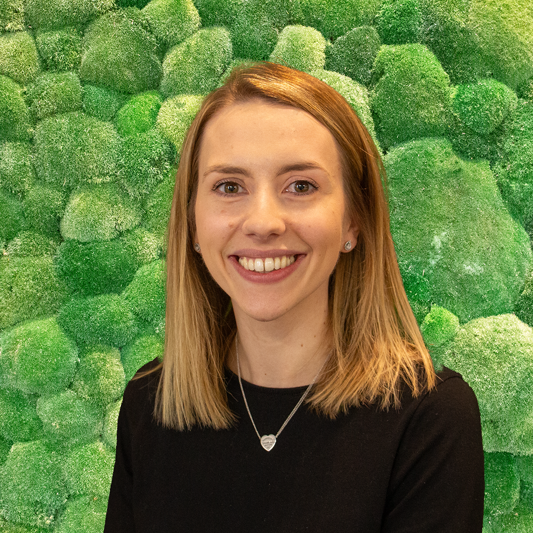 Headshot of a smiling young blonde woman in a black top against a vibrant green textured moss wall.