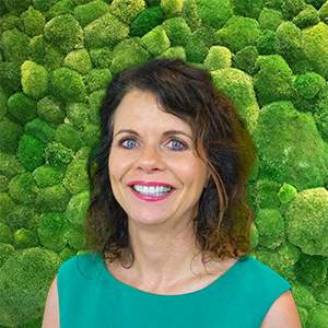 Headshot of a smiling woman with dark hair, blue eyes, wearing green, against a green moss background.