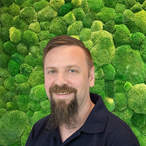 Smiling man with beard in a dark polo shirt against a textured green moss wall, ready to help.
