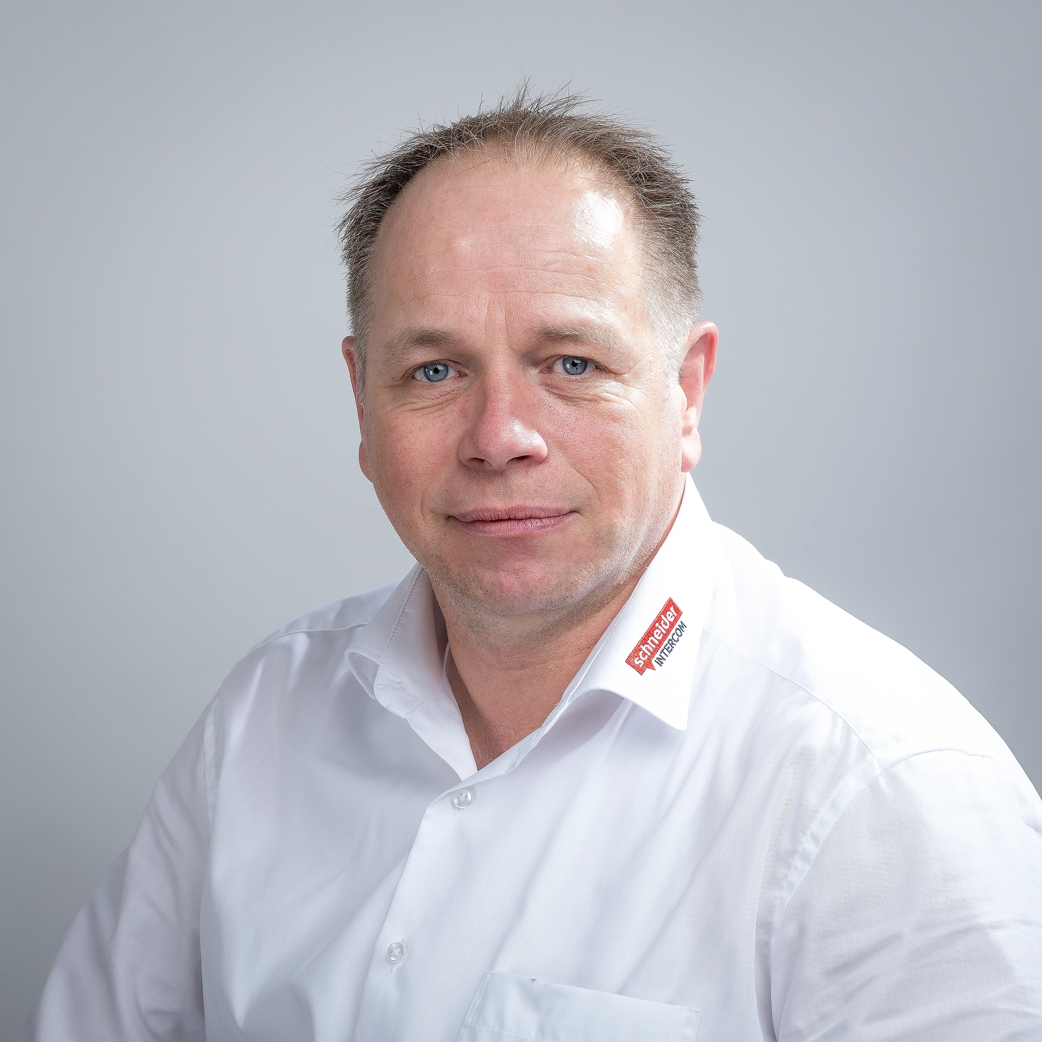 Mann in weißem Hemd mit Schneider Intercom Logo lächelt. Experte für Kommunikationslösungen.