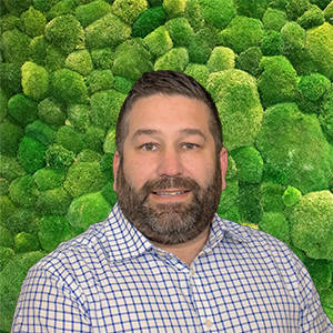 Headshot of a smiling man with a beard and checkered shirt against a green moss-textured wall.