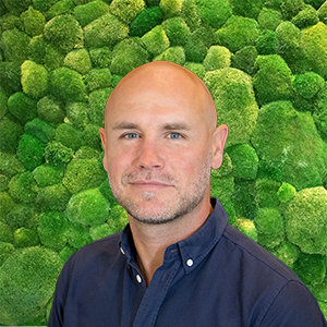 Portrait of a smiling man with a beard and blue shirt against a green moss wall.