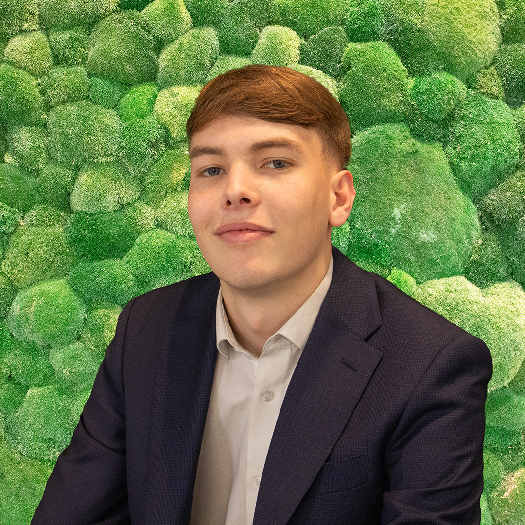 A young man in a blue suit smiles in front of a green moss wall.