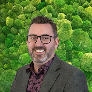 Headshot of a smiling man with glasses and a beard against a green moss background.