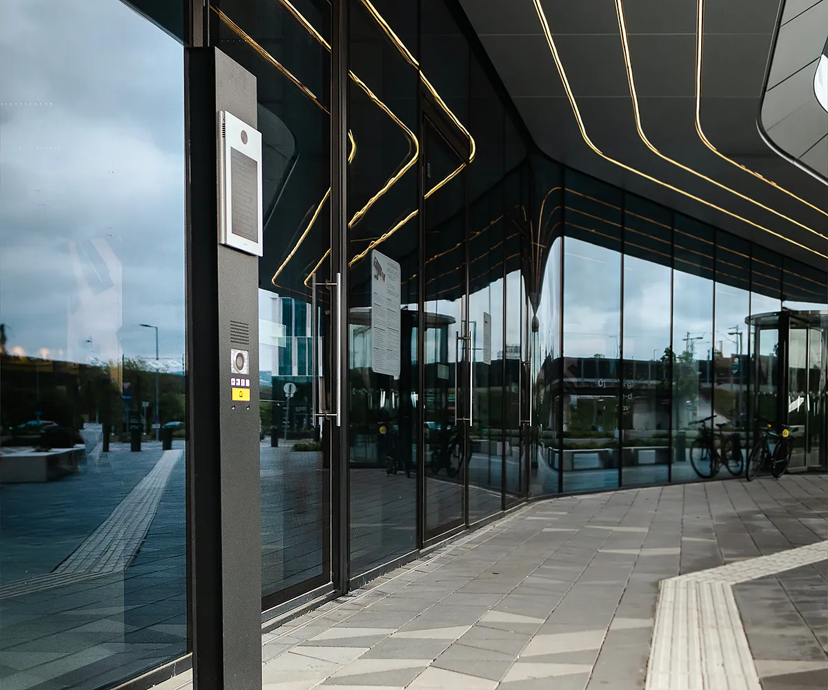 Modern kaputelefon sötét oszlopon üveg épületbejáratnál. Fénycsíkok és utca tükröződései.