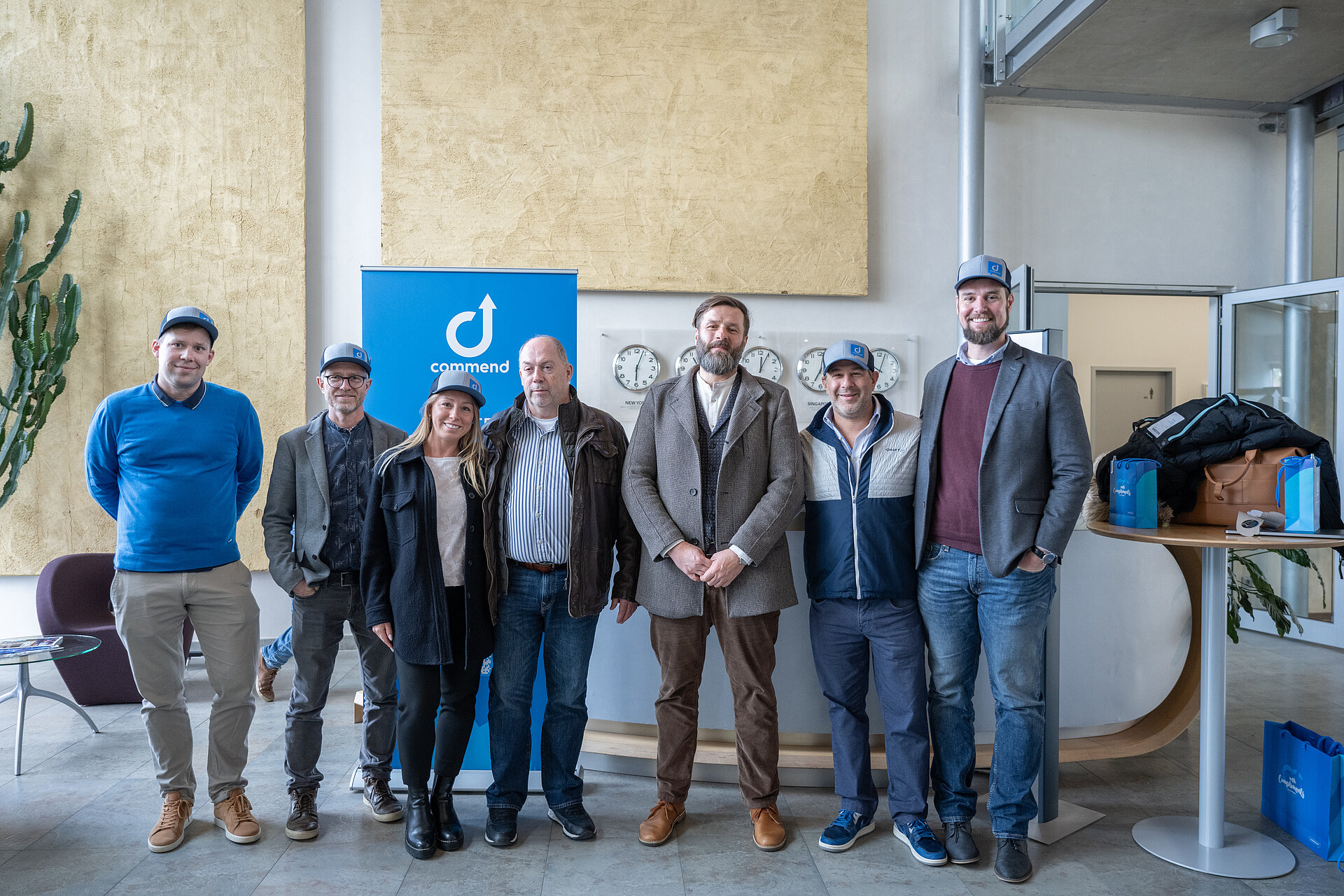 Seven people in Commend caps standing in an office with a blue logo banner, showcasing our communication solutions.