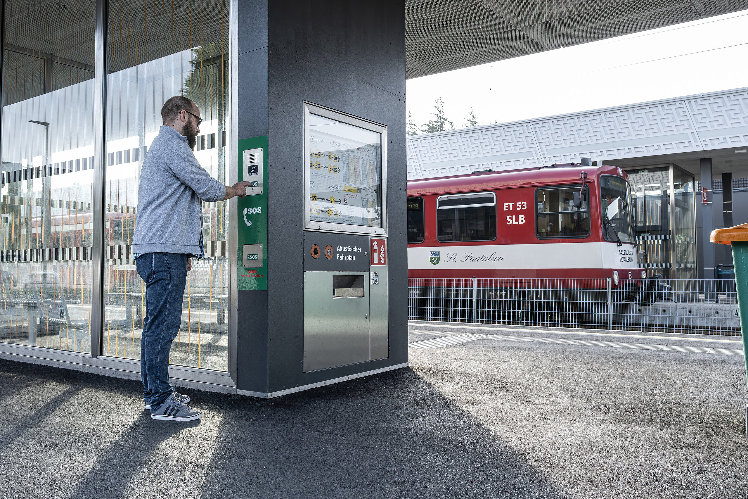 Uomo su un binario della stazione preme un pulsante SOS su un interfonico vicino a un orario acustico.