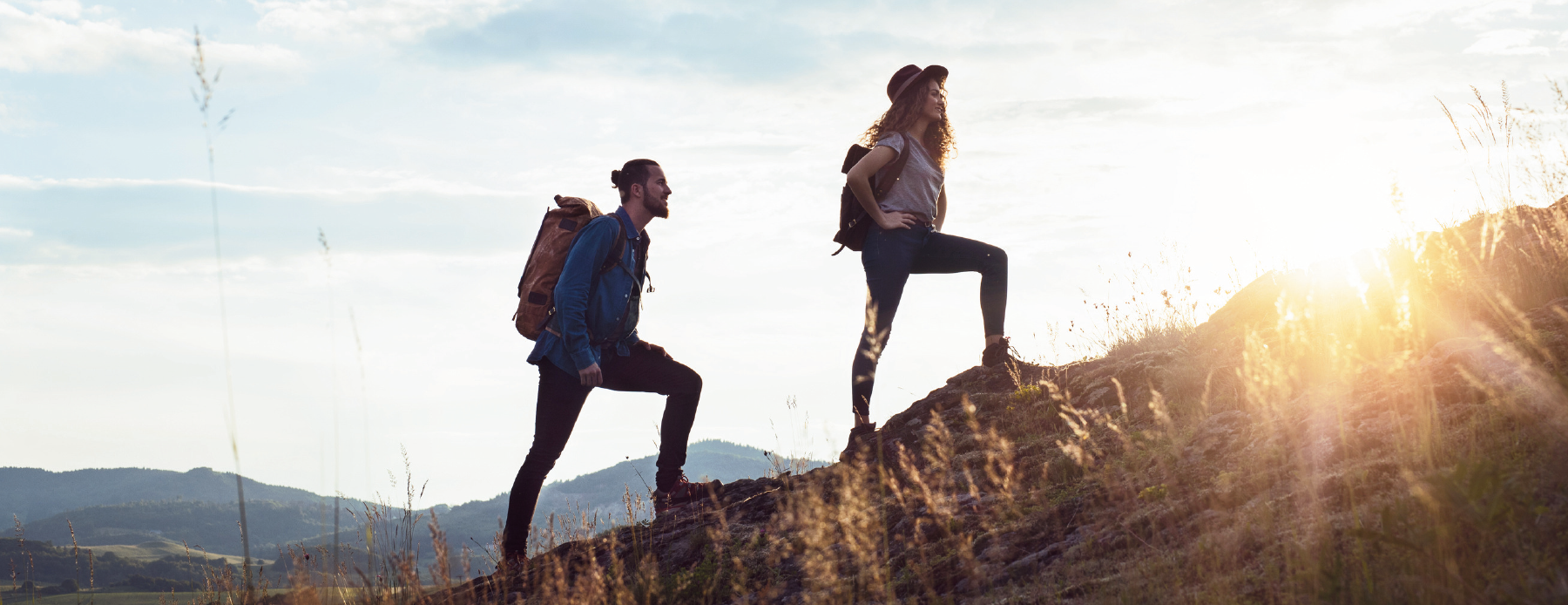Two hikers with backpacks ascend a rocky hill at sunset, silhouetted against a bright sky.