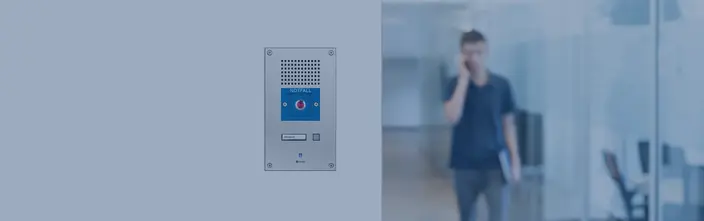 Panel de interfonía plateado con botón de emergencia rojo ('NOTFALL') y etiqueta 'Secretaría' en oficina, persona borrosa en segundo plano.