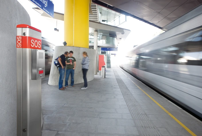 SOS Intercom at a Modern Station Silver and red SOS intercom on a station platform with a blurry train passing by and people in the background.