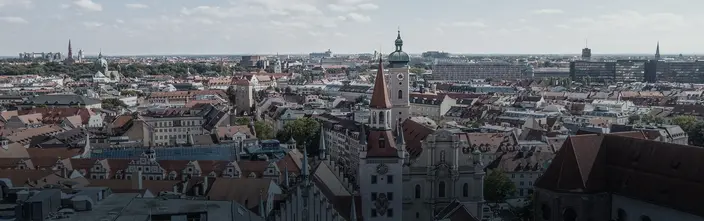 Munich Cityscape: Rooftops and Spires Aerial panoramic view of Munich city, showing red-tiled roofs, church spires, and a distant modern skyline.
