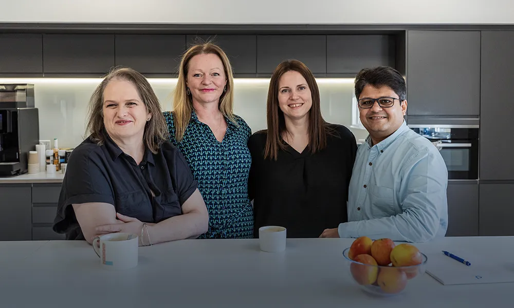 Team Collaboration in the Office Kitchen Four smiling colleagues in an office kitchen, standing behind a counter with mugs and apples.