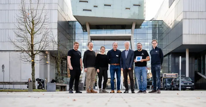 Team gathered outside modern building Seven professionals posing outdoors in front of a modern glass building, one holding a document with a 'd' logo.