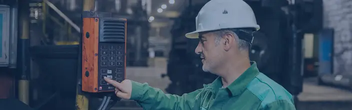 Man in hard hat using an orange industrial intercom in a factory. Keypad and speaker visible.