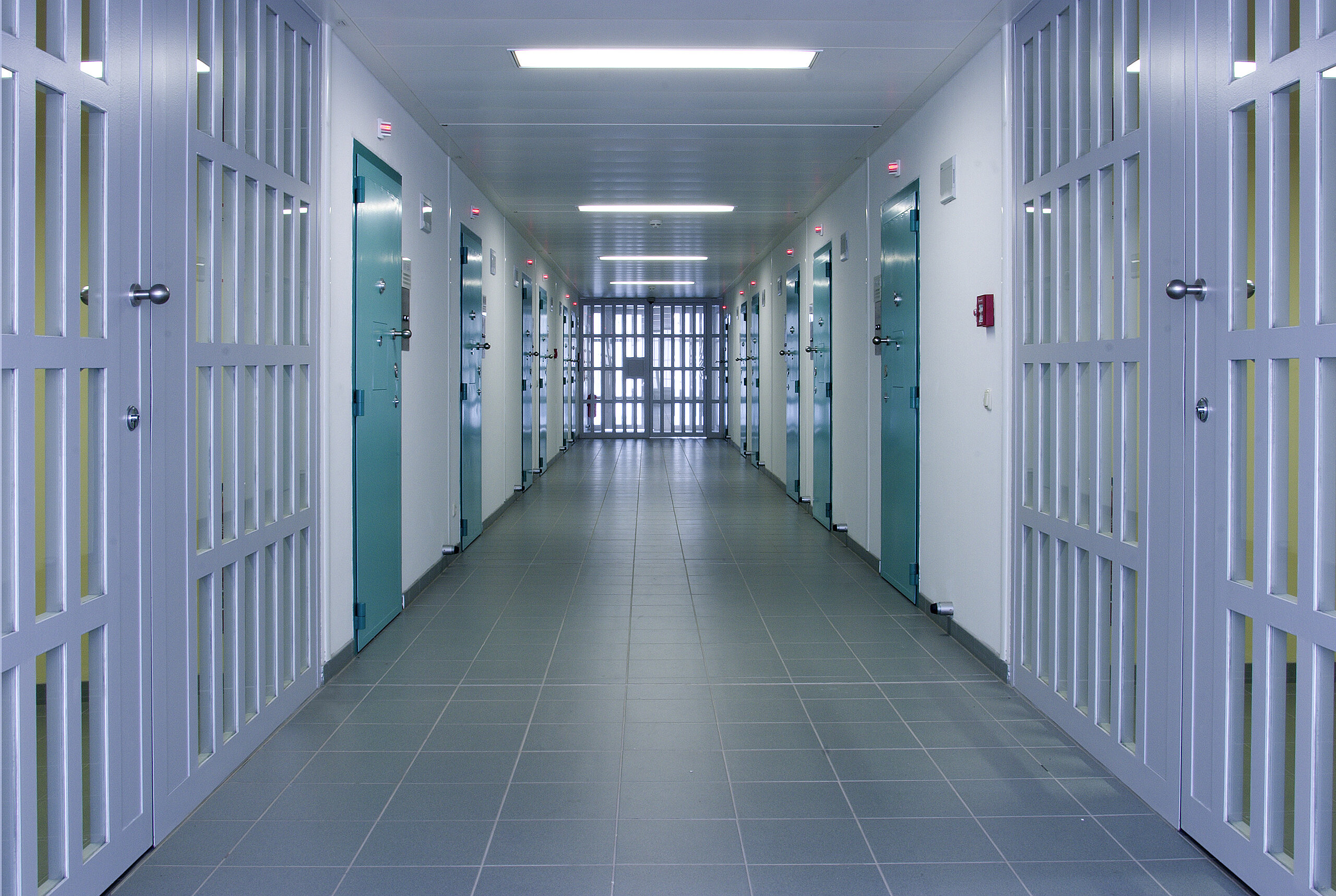 Lungo corridoio sterile in struttura sicura con molte porte turchesi e cancello sbarrato.