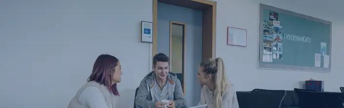 Three young people talking in a common area with an intercom system and a bulletin board visible.
