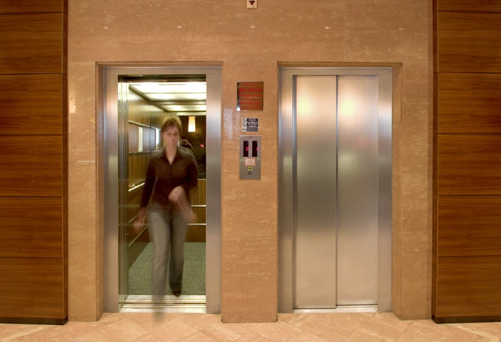 Woman exiting an open elevator in a lobby. An intercom panel is visible between two elevator doors.