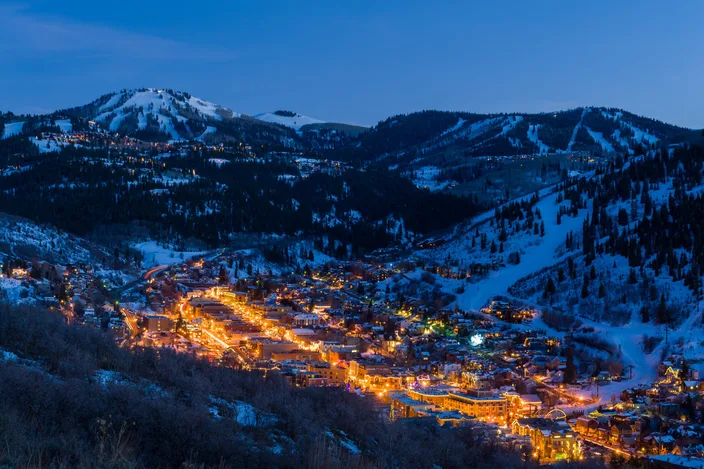 Park City bei Dämmerung im Winter Luftaufnahme einer hell beleuchteten Stadt in einem schneebedeckten Bergtal mit Skipisten bei Nacht.